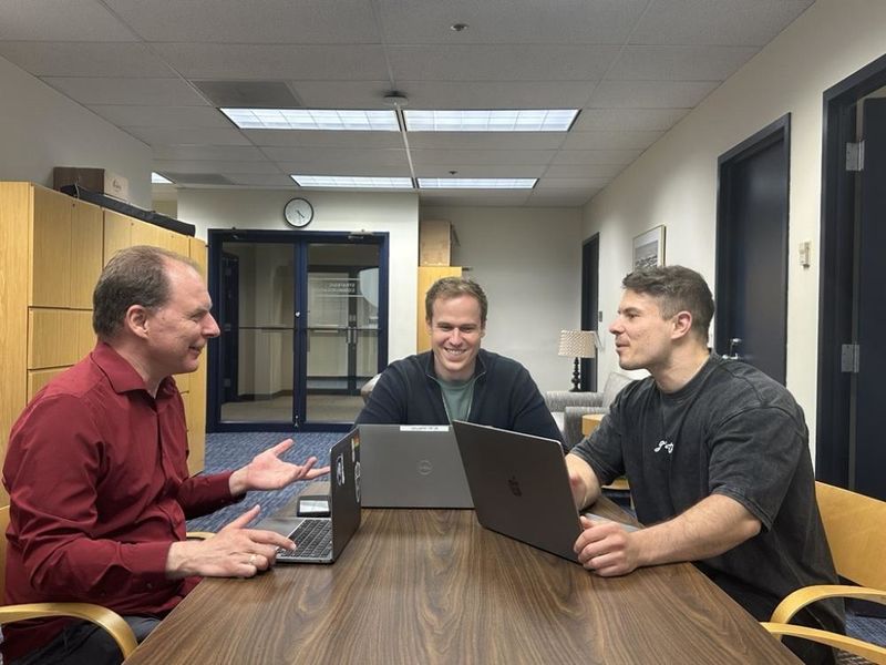Three men sit at a table with their laptops in an office, talking about their work. 